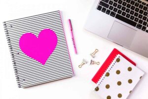 Desk with a notebook with a pink heart on it, a pen, and the edge of a laptop
