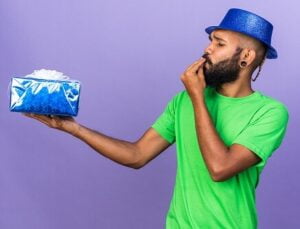Bearded African male in a green tshirt and blue hat holding out a gift wrapped in blue paper with a purple background