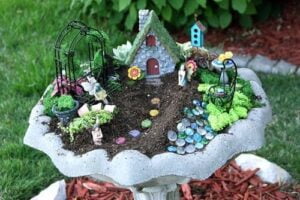 outdoor fairy garden with house, pathway, fence and plants in a bird bath