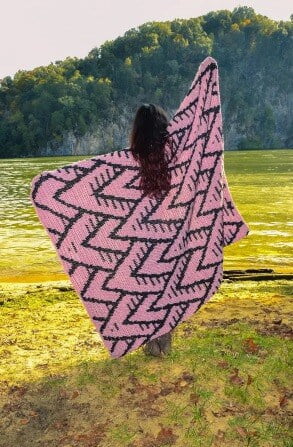 blanket in pink and black crocheted using the tapestry technique is being held up by a dark haired woman in a filed surrounded by trees