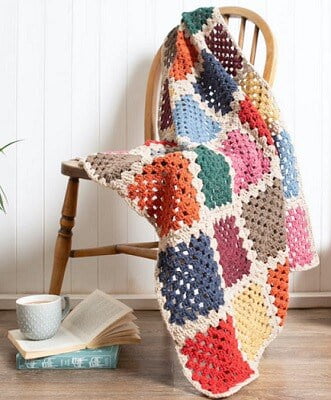 blanket crocheted in red, blue, orange, pink and mauve is draped over a chair with a pile of books and a coffee cup on the floor