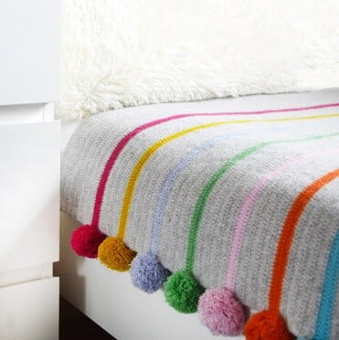 Crochet blanket in grey with orange, pink, blue, purple stripes with a pom pom on the end of each colour stripe