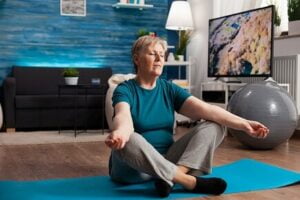 older white woman wearing a blue top and grey trousers is sitting on the floor in a front room meditating
