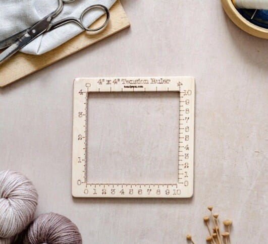 A tension ruler is laid flat with scissors and yarn to show it's for knitting and crocheting
