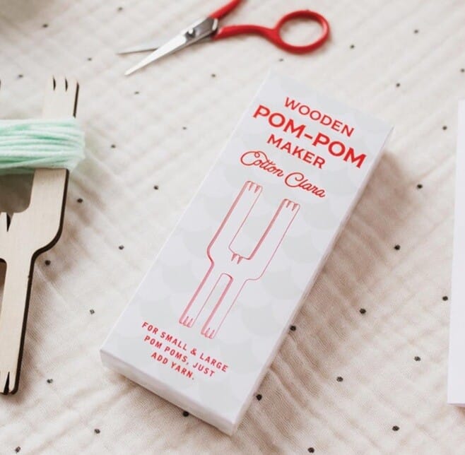 wooden pom pom maker is displayed on a table alongside the packaging and scissors