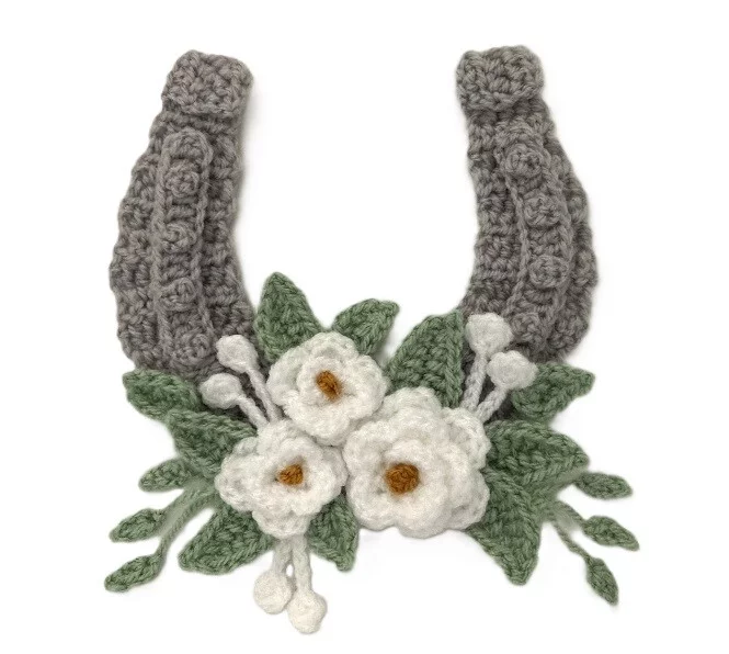 A horseshoe is laid flat. It is crocheted in an olive colour with a spray of crocheted roses at the bottom