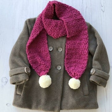 Flat lay of a child's coat with a purple crochet scarf with white pompoms on the ends