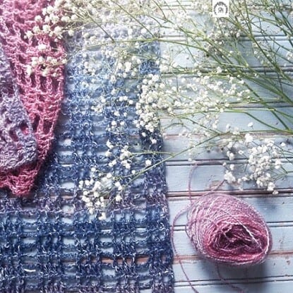 A lacy crochet scarf in blue and light purple is on a flat surface with a spray of white flowers and the yarn used 