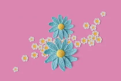 2 large blue crochet daisies surrounded by smaller white daisies on a pink background