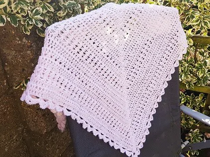 Light pink crochet shawl with a delicate lacy texture and scalloped edge, draped over the back of a black outdoor chair. The triangle-shaped shawl features an openwork stitch pattern, creating a soft and airy look