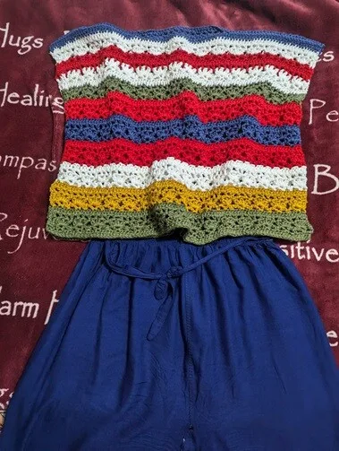 This image shows a handmade crochet top laid out on a dark red blanket with positive words like "Healing," "Hugs," and "Compassion" written on it. The top features wavy horizontal stripes in alternating colors—blue, white, red, green, mustard yellow, and olive—paired with a flowy pair of high-waisted blue pants tied at the waist.