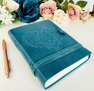 leather journal with a blue colour, a pen is next to the journal and blue and pink roses at the top of the image