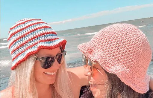Two women are smiling at each other at the beach wearing sun hats