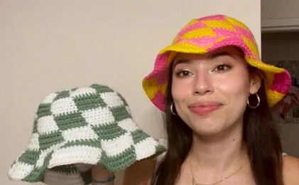 A woman is wearing a checkered summer hat in pink and yellow while holding another hat in green and white