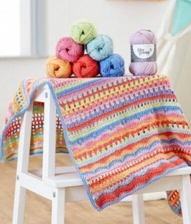 A colour crochet blanket is draped over a chair with the balls of yarn on top of the blanket