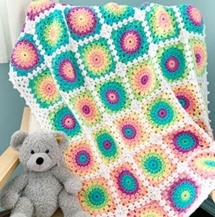 sunburst granny square blanket crocheted in the colours of the rainbow is draped over a chair with a teddy sat on the seat