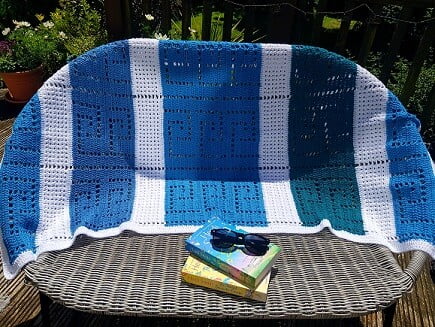 crochet blanket in blues and white draped on the back of garden sofa with two books and sunglasses