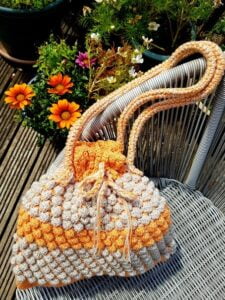 crocheted bucket bag in orange and oatmeal colours is on the seat of garden furniture with orange coloured flowers in the background