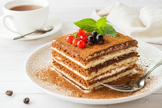 A slice of coffee tiramisu on a white plate with a fork, a cup of coffee and coffee beans on the table