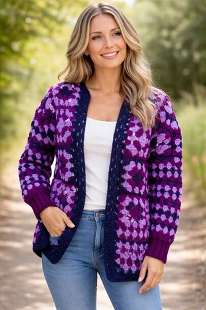 A woman is wearing a granny square cardigan in pink and purple. She is standing with a hand in her jeans pocket