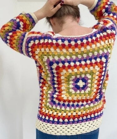 A woman is facing away with her arms holding up her hair and she is wearing a granny square crocheted sweater