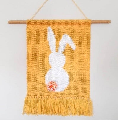 the back of a white bunny with a fluffy tail on a background of orange on a crocheted wall hanging