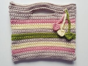 crocheted tote bag in different coloured stripes with a crocheted heart bag charm on the right hand side