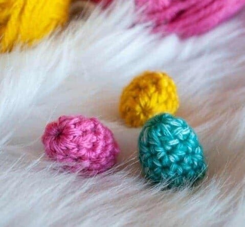 3 mini crochet Easter eggs in pink, turquoise and yellow are sitting on a furry cover