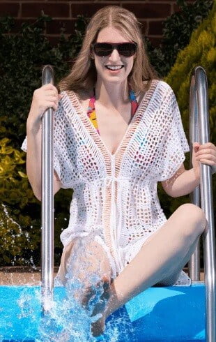 A woman is sitting by the side of a pool splashing her feet. she is wearing a crochet cover up