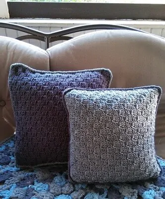 A dark grey and light grey crocheted pillow is nestled on a sofa