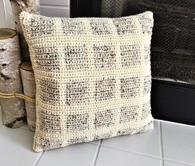 Pillow with a cream background and grey squares is placed on the hearth of a fireplace