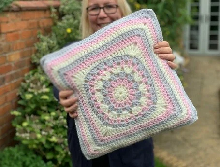 A woman is standing and holding up a large granny square pillow