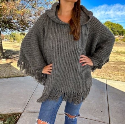 A young woman is standing on a path with her hands on her hips wearing ripped jeans and a hooded crochet poncho
