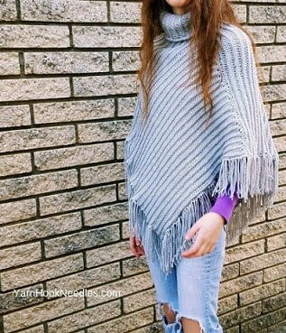 A woman is standing in front of a brick wall wearing ripped jeans and a grey poncho
