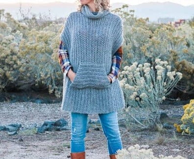 A woman is standing in front of trees wearing a grey cowl necked poncho