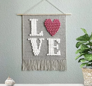 Crochet Wall Decor with the word 'love' crocheted in bobble stitch is hung on a grey wall with a plant on one side