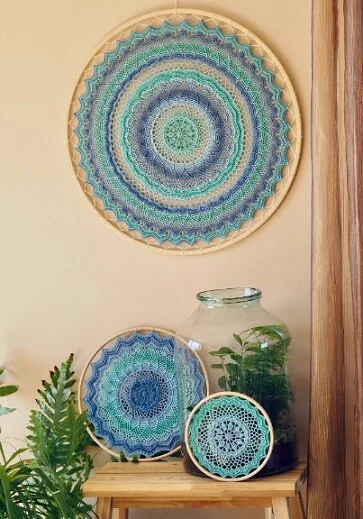 One large size mandala in blue yarn is hung on a wall and two smaller sized wall hangings are resting below the larger one