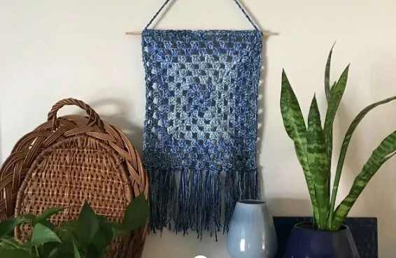 A classic granny square with a fringe is hung on a wall beside a wicker tray and a plant
