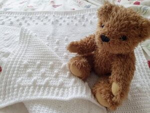 A crochet blanket is folded over with a teddy bear sat on the corner
