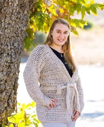 A woman is standing next to a tree and facing sideways styling a belted chunky crochet cardigan