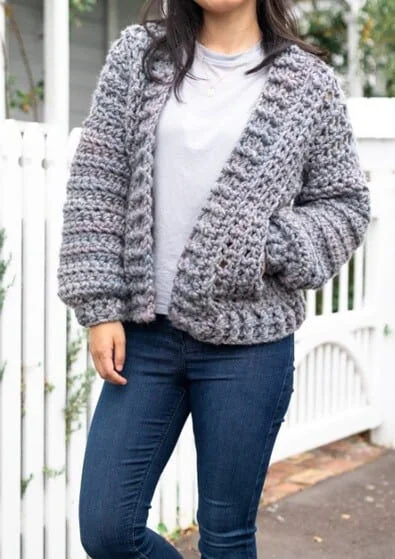 A woman is wearing a grey crocheted bomber jacket with one hand in a pocket