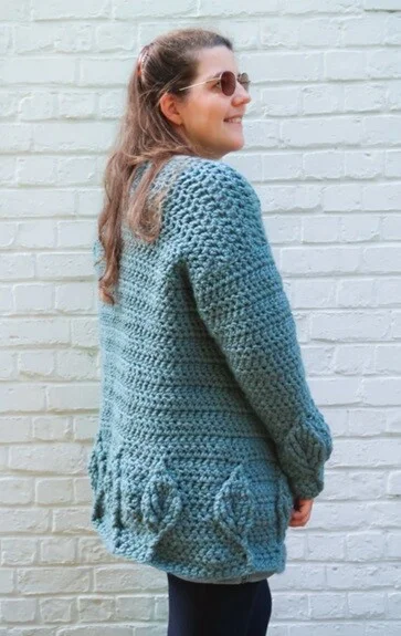 A woman is wearing a chunky cardigan with textured leaves on the sleeves, pockets and hem