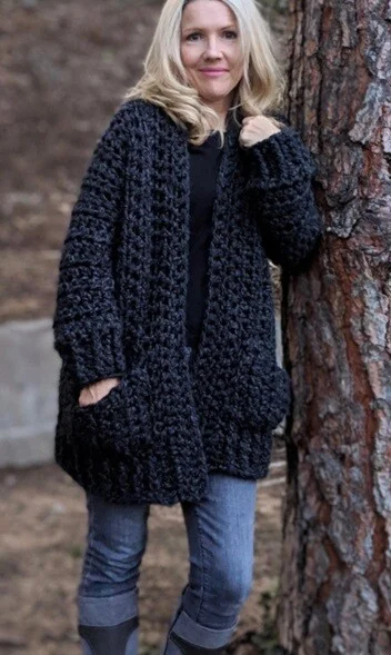A woman is leaning against a tree.  She is wearing jeans, boots and a black oversized crochet cardigan