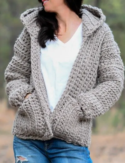 A woman is standing in the woods wearing jeans and chunky crocheted hoodie
