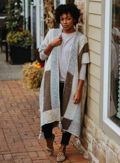 A woman is leaning against a wall wearing leggings and a long patchwork cardigan