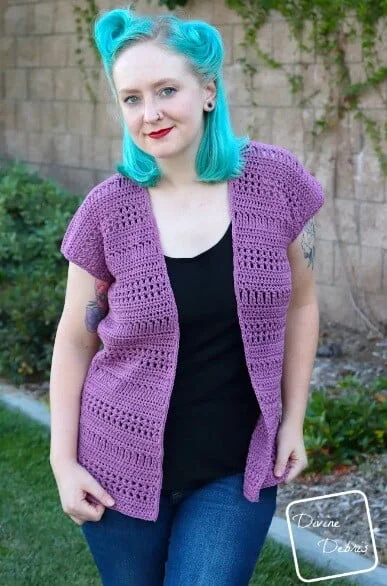Woman with turquoise hair modeling a short-sleeved crochet summer cardigan in plum purple with horizontal openwork stripes, worn over a black top and jeans, standing outdoors near a garden wall.