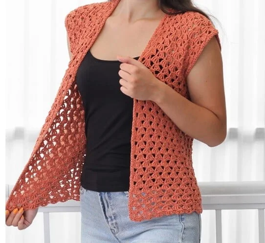 Woman wearing a rust-colored crochet summer cardigan with cap sleeves and an airy, shell-stitch pattern, styled over a black tank top and light blue jeans.