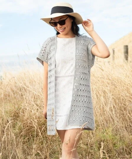 Woman standing in a field wearing a light grey crochet summer cardigan with short sleeves and an open front over a white dress, accessorized with a sunhat and sunglasses.