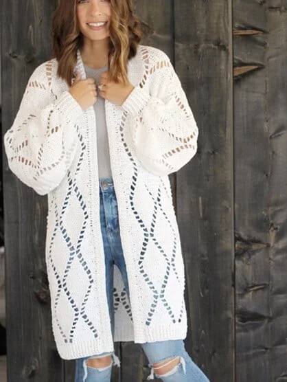 Woman wearing a long white crochet cardigan with oversized sleeves and diamond-shaped openwork patterns, styled over a white top and distressed jeans, standing in front of a rustic wooden wall.