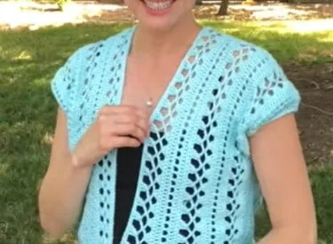 Smiling woman outdoors wearing a light blue crochet summer cardigan with cap sleeves and an open front, featuring vertical lace-style stitches in a leafy pattern over a black top.